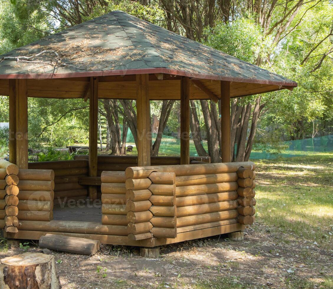 An open gazebo made of round logs in the center of the park, with branches and foliage on the roof. photo