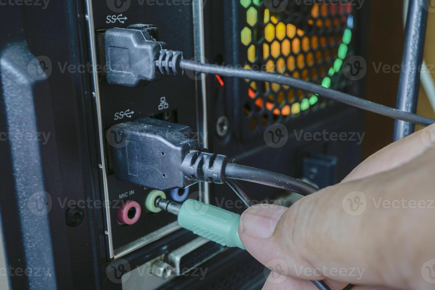 Hand connects green audio cable to microphone input on computer back panel, alongside USB and HDMI ports. setup is part of desktop computer with visible cooling fan photo