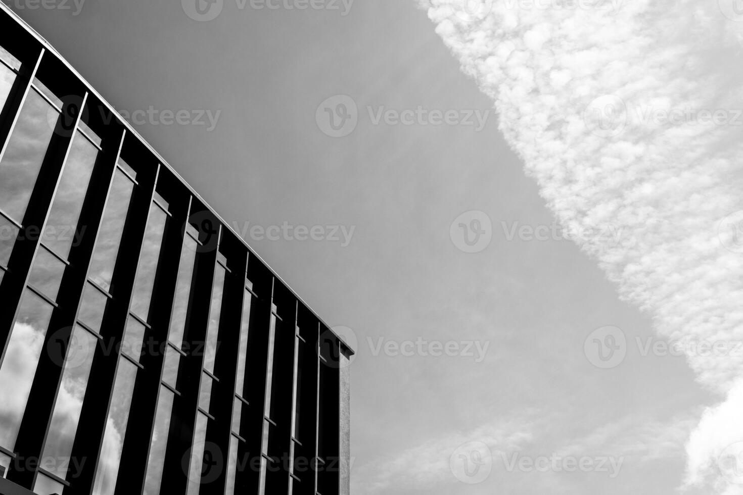 Black and white photo of a modern building facade with vertical lines and glass windows against a sky with textured clouds. Suitable for illustrating architecture, urban design, or minimalism concepts in editorial or commercial projects.