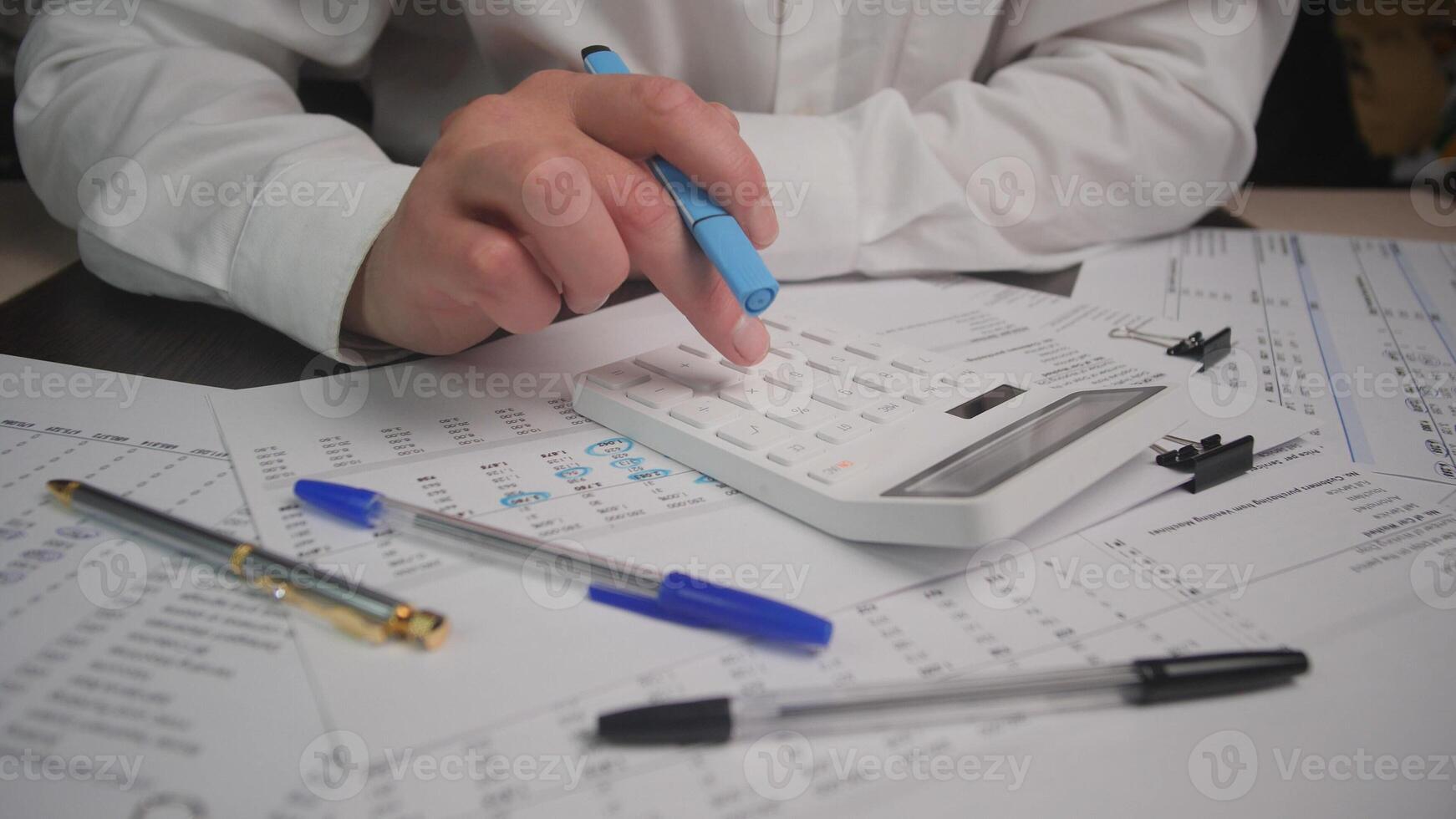 A person is using a calculator to manage finances while sitting at a desk filled with paperwork and various pens, focused on budgeting tasks in an office. photo