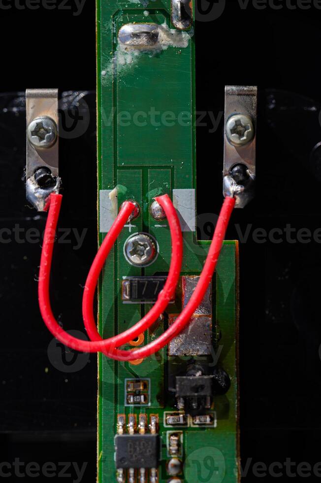 A detailed, vertical macro shot of a green printed circuit board PCB with a red wire soldered to two points. The image captures electronic components and intricate circuitry details. photo
