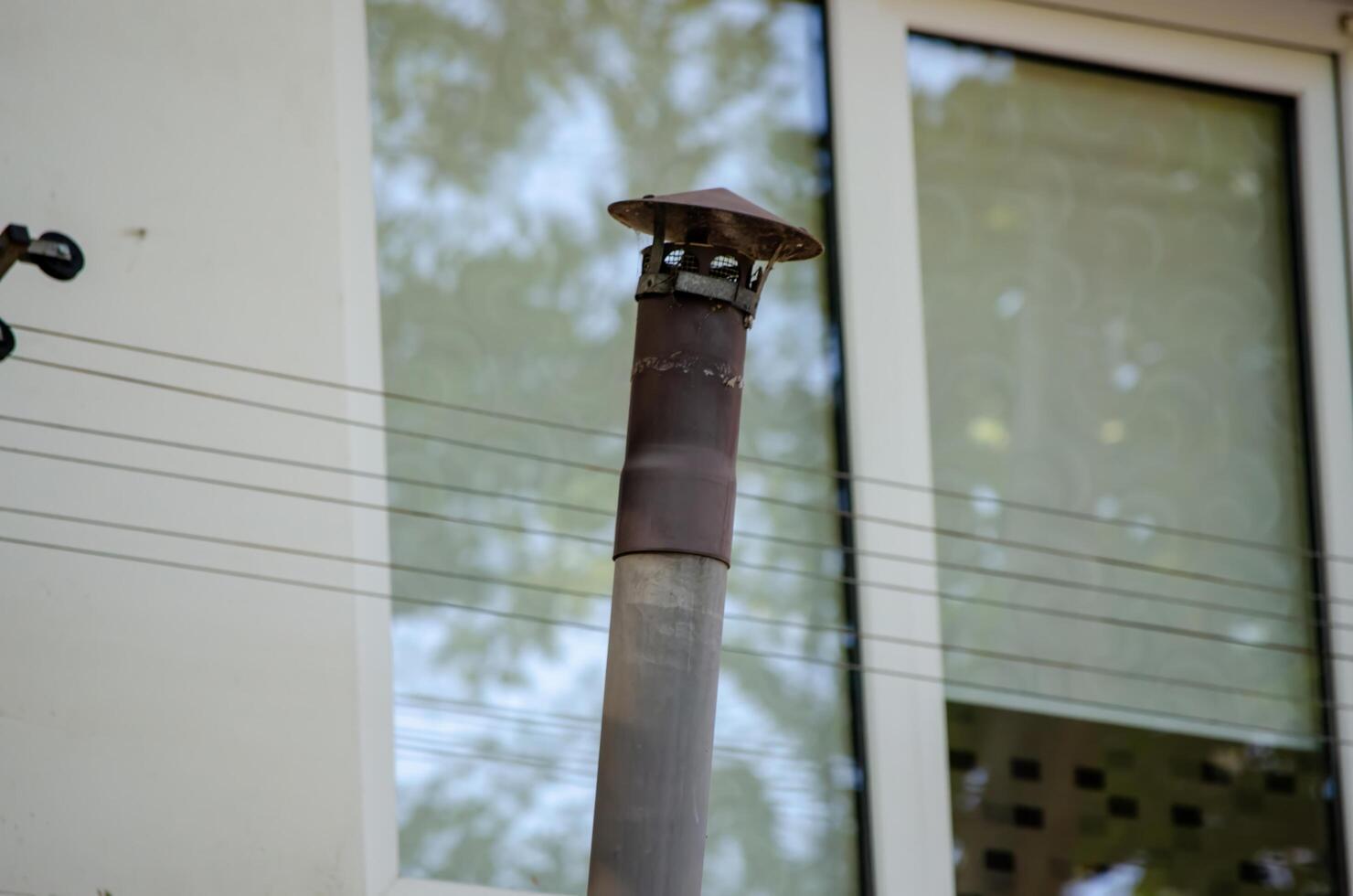 A rusty pipe sticking out of a building. The pipe is covered in rust and has a metal cap on top. The building is surrounded by wires and has a window with a patterned curtain photo