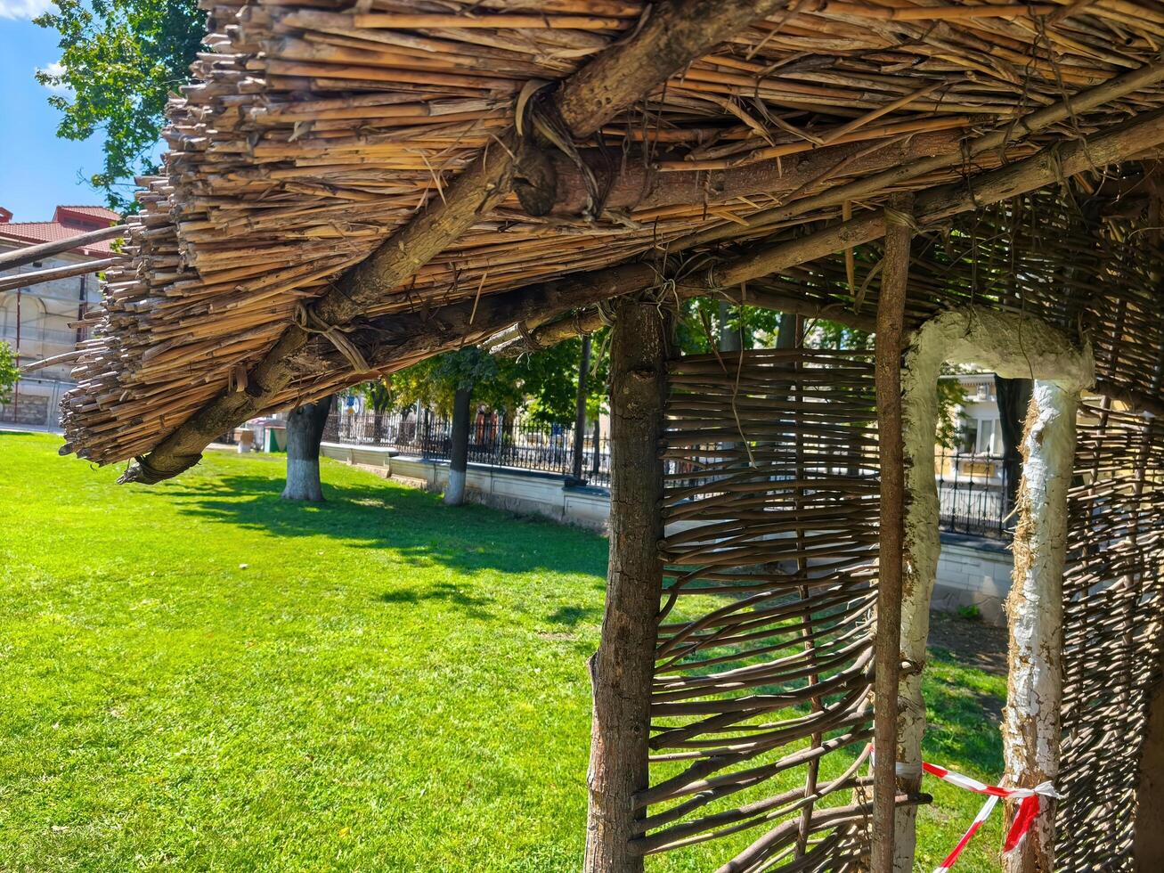 A wooden structure with a thatched roof sits in a grassy field. The structure is surrounded by trees and he is a part of a park photo