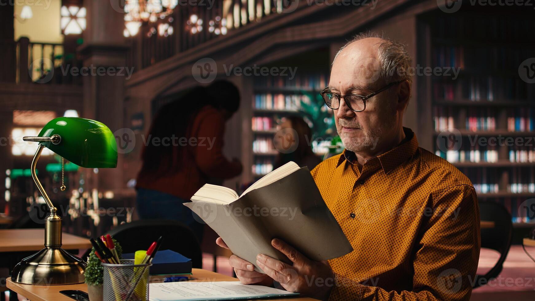 Old person with spectacles enjoys reading a book at the library, being happy and enjoying recreation. Aged man with book reading hobby, retirement leisure activity in quiet space. photo