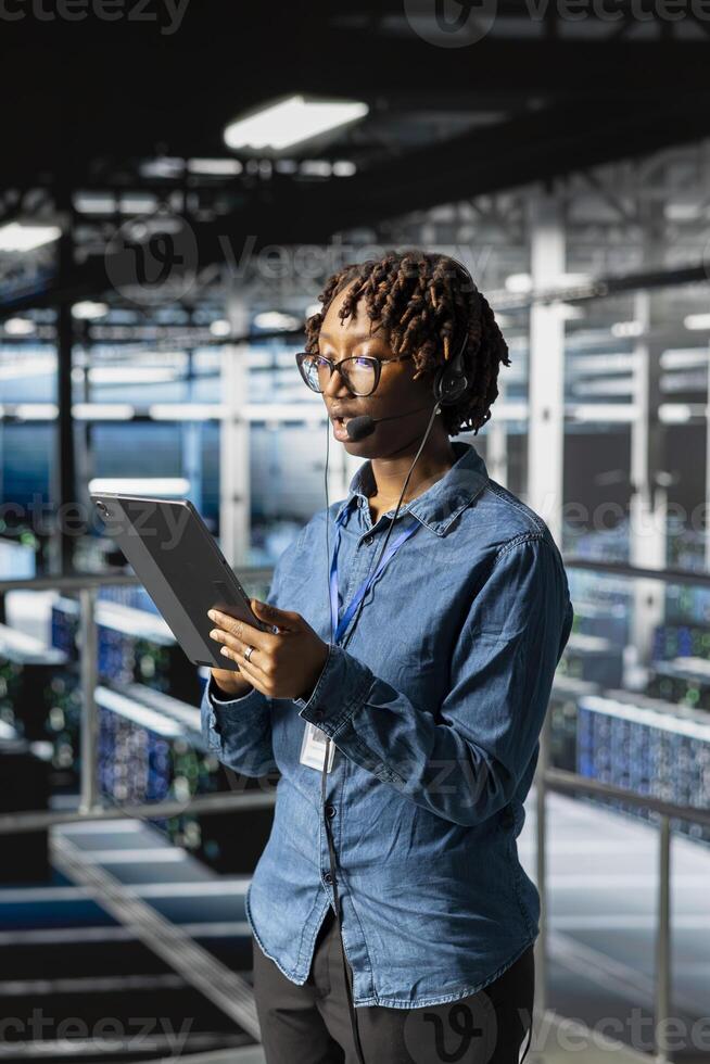 Data center specialist with headset microphone for remote diagnostics on tablet, industrial platform. Technician monitors and guides users through infrastructure troubleshooting steps. photo