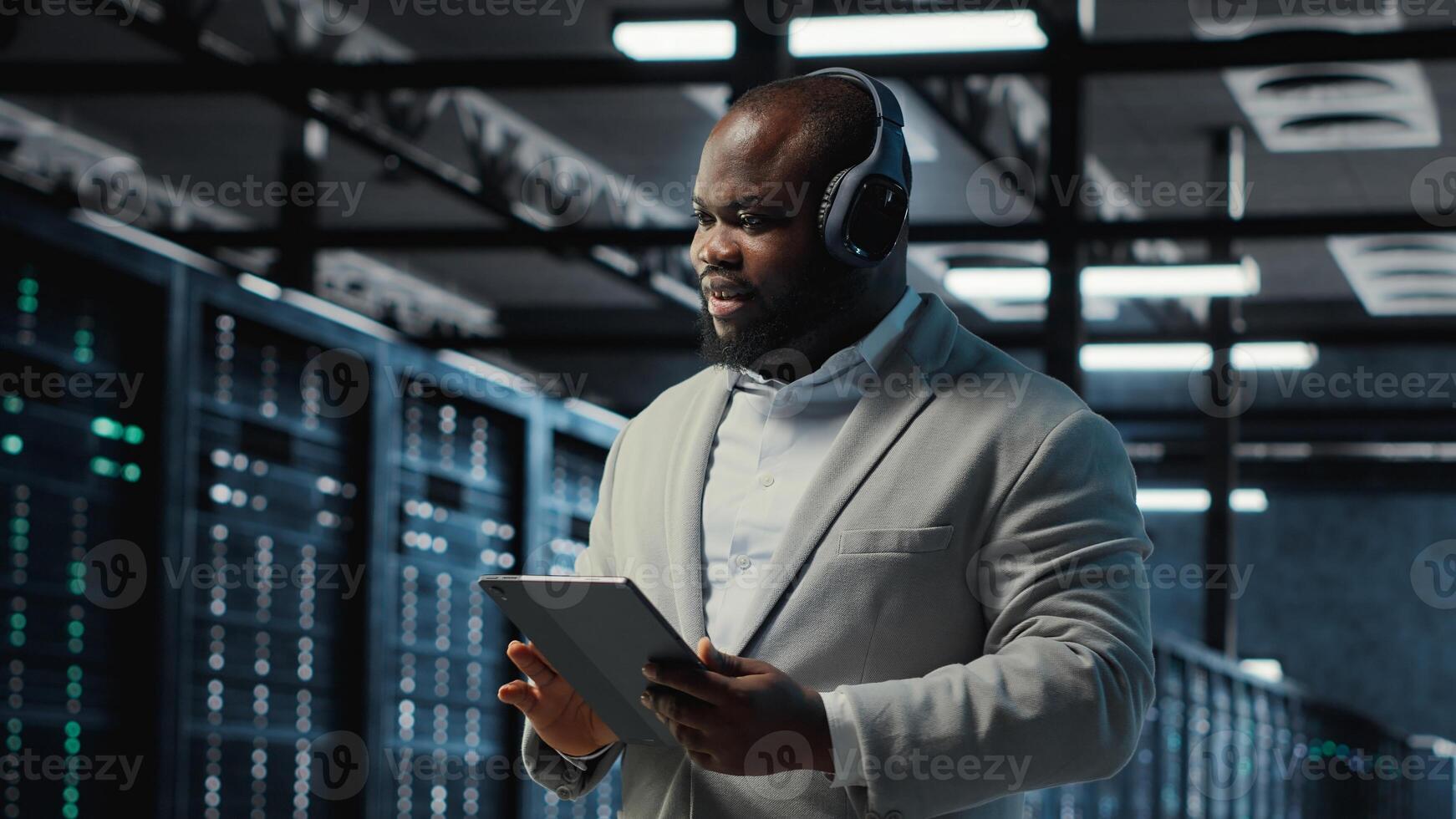 Server room programmer wearing headphones troubleshooting errors using software on tablet. IT specialist listening music, walking in data center fixing bugs using device , camera B photo