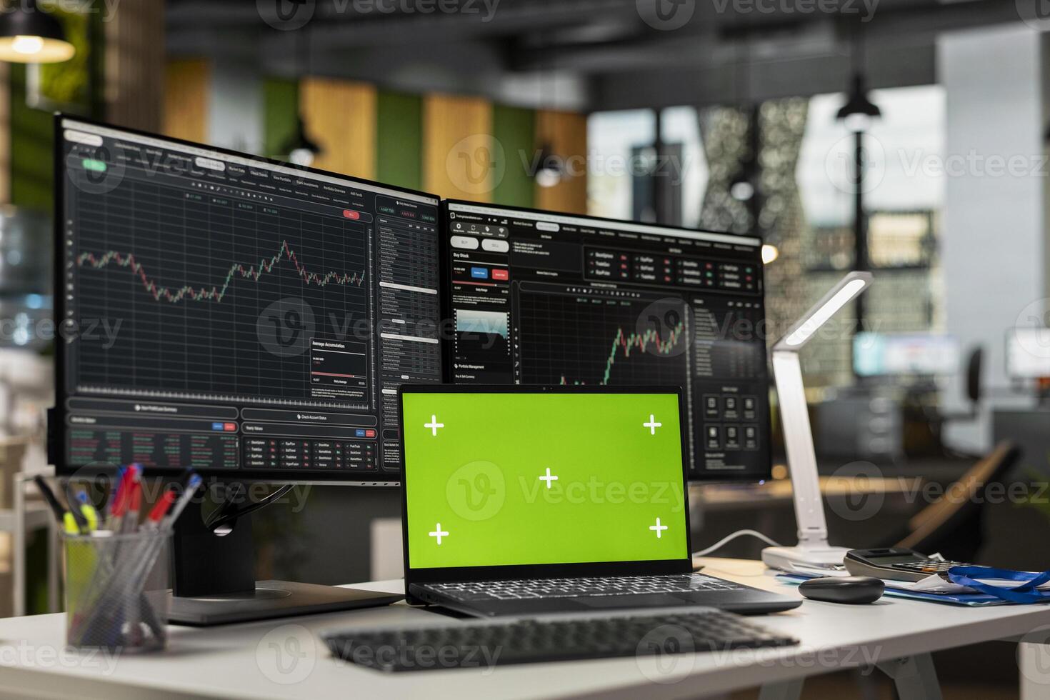 Green screen laptop in brokerage office next to workstation monitor displaying live stock charts. Mockup notebook on desk and computer with financial software in prop firm workspace photo