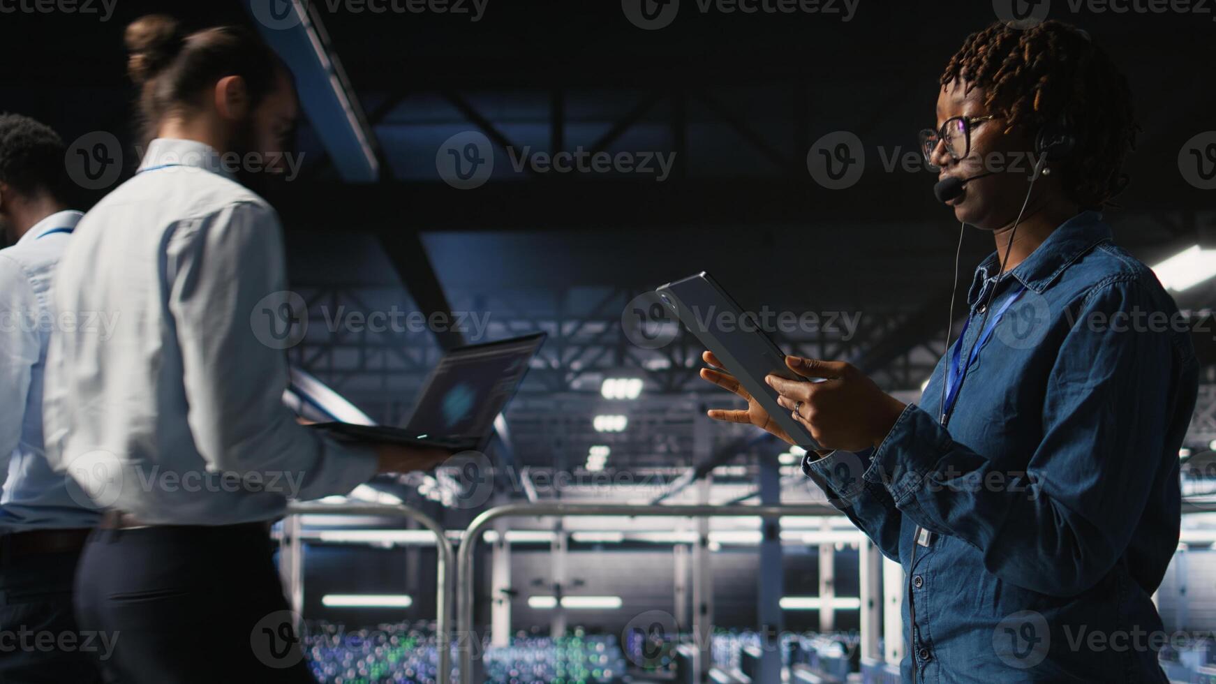 Server room admin using tablet and headset microphone to provide infrastructure tech support. Data center IT specialist resolves backend issues while coordinating with remote teams, photo