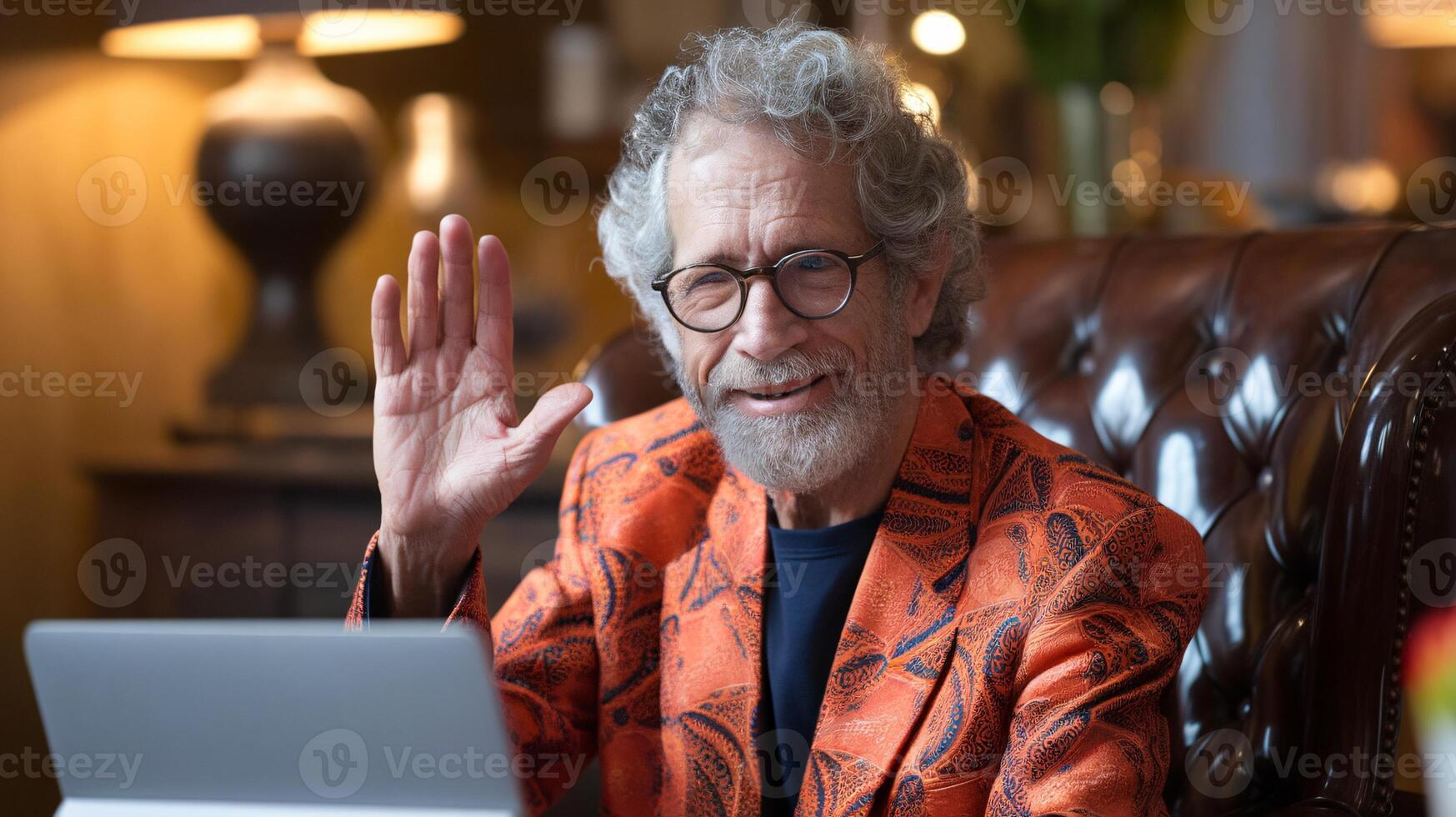 Smiling elderly man in an orange patterned blazer waves while using a tablet in a cozy, stylish setting. photo