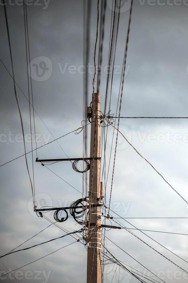 Tangled Wires On A Concrete Pole. A low-angle shot of a concrete utility pole with a chaotic tangle of electrical and communication wires against a cloudy sky. photo