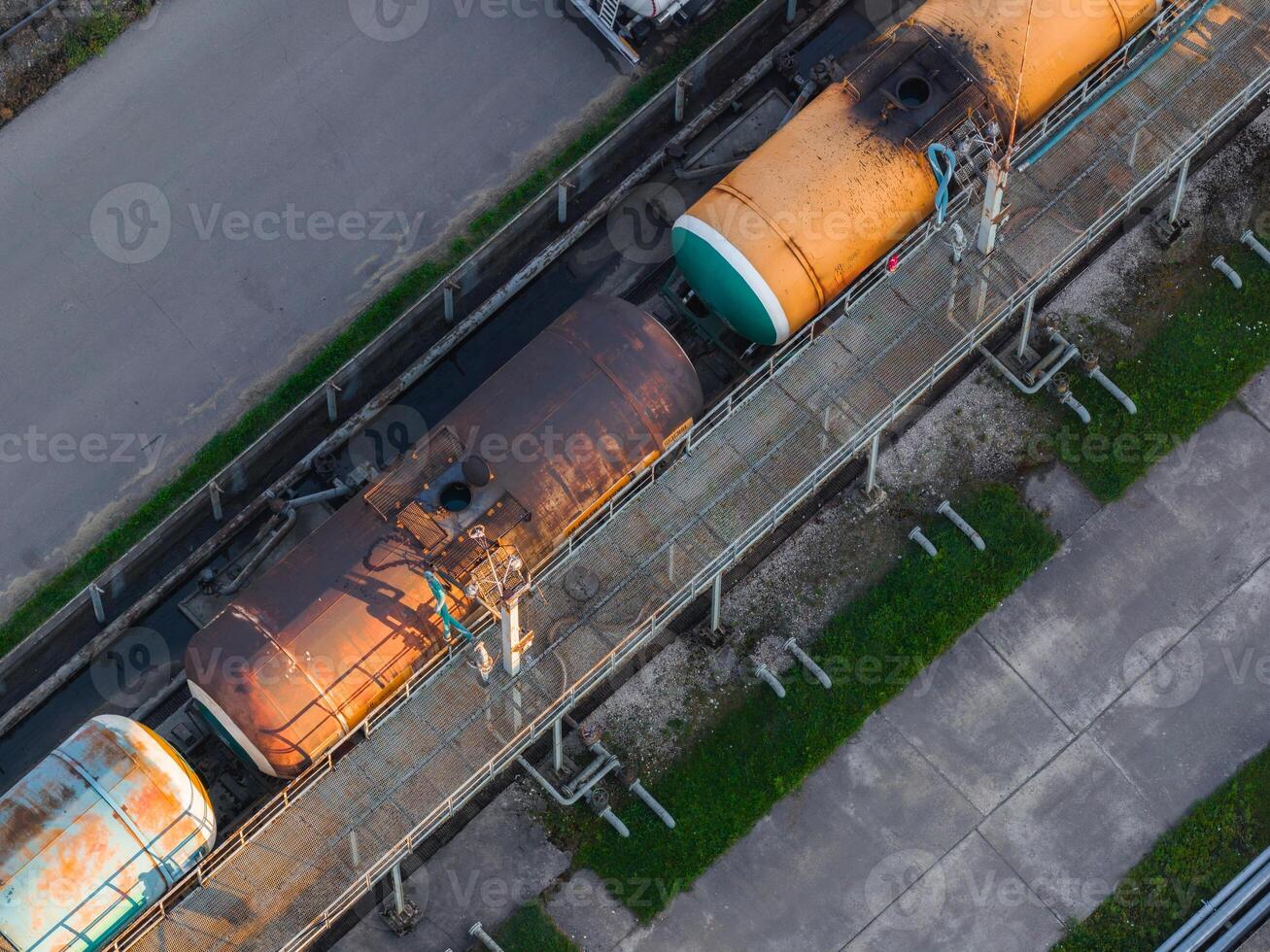 Aerial view of colorful and rusted tanker railcars on parallel tracks in Riga, Latvia, bordered by a road and grassy patches with visible infrastructure. photo