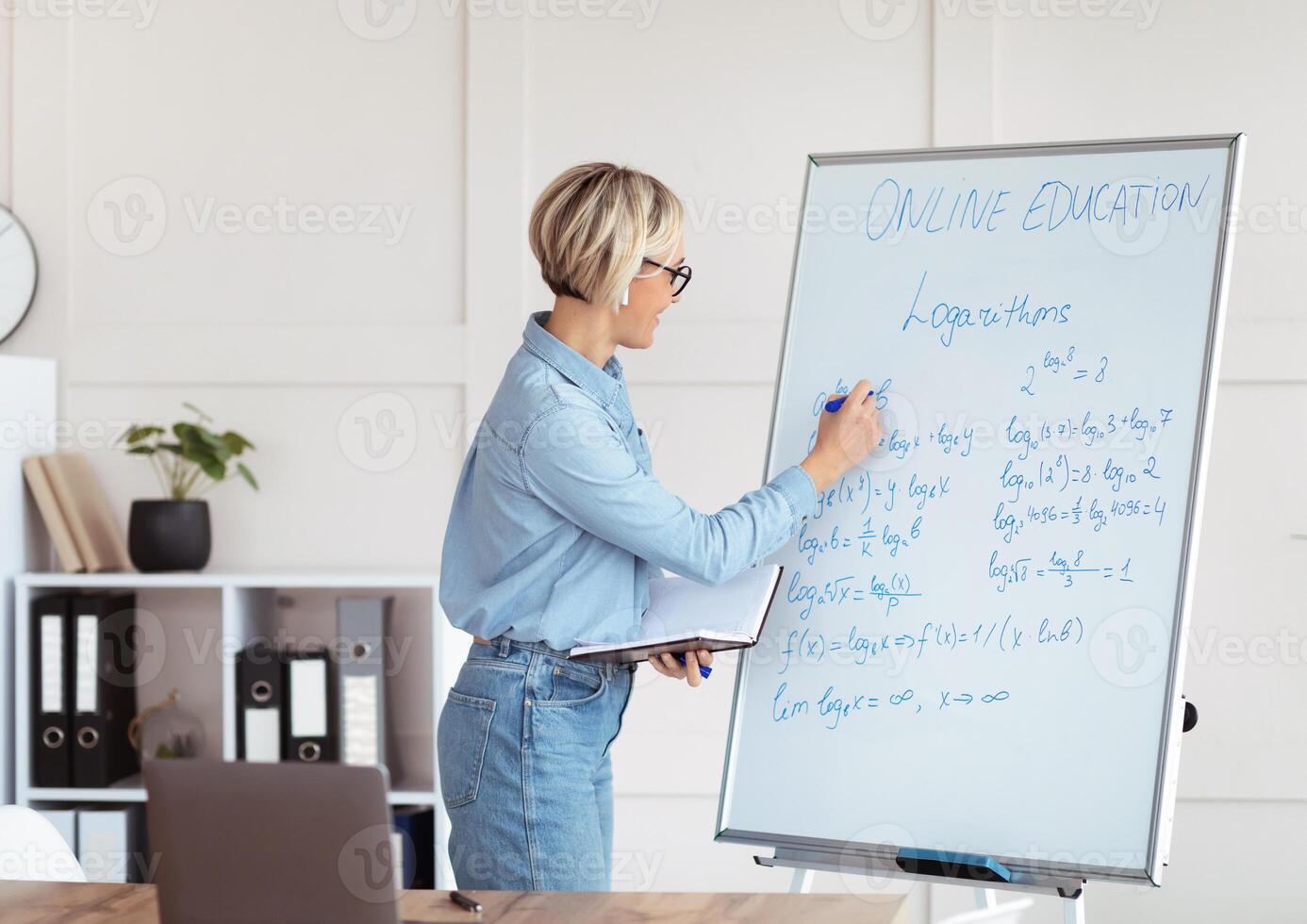 Female university tutor solving complicated math sums during online lesson, explaining logarithms on laptop from home. College professor teaching calculus, giving remote lecture photo