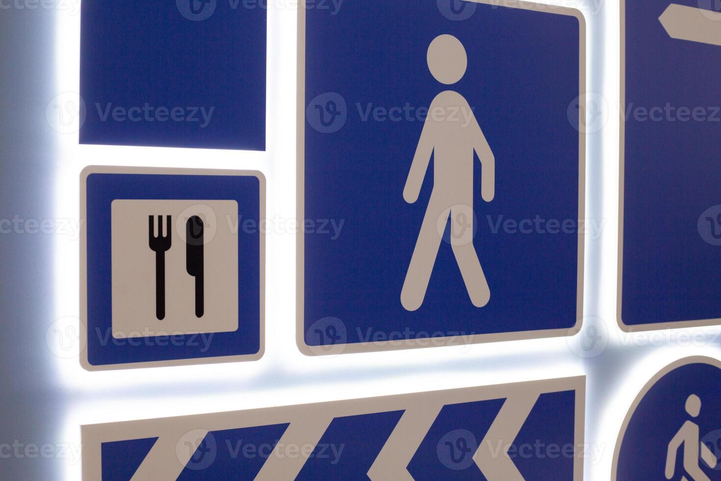 Various directional and informational signs. Symbols include pedestrian icon, dining symbol with utensils, and directional arrow, all set on blue background with illuminated edges photo
