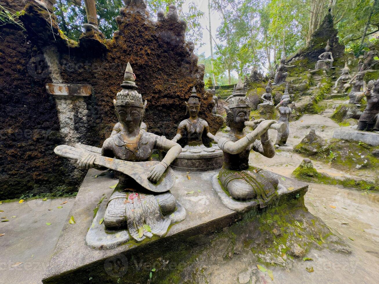 A close-up, medium-shot view of ancient stone statues of mythical musicians, one playing a stringed instrument and another holding a flute-like object, situated in a lush, moss-covered garden. The photo