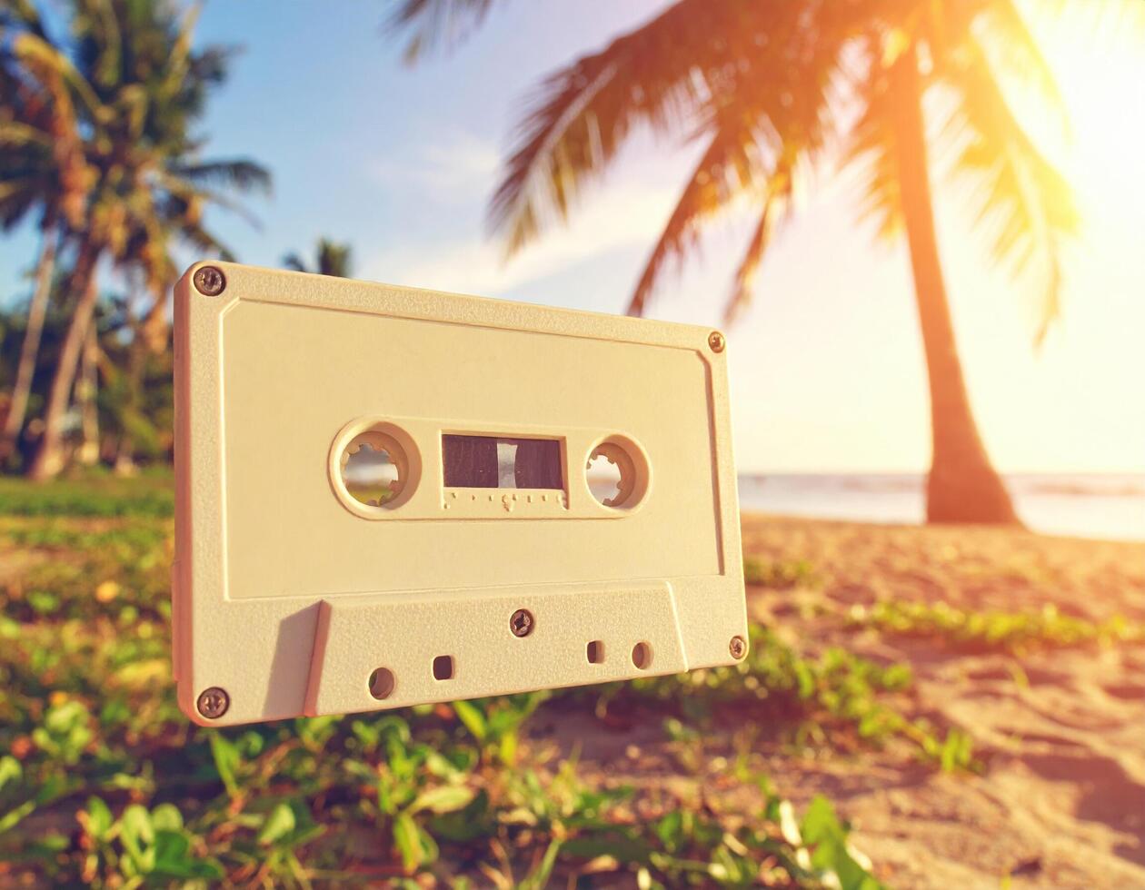 Retro cassette tape floats in air against tropical beach backdrop, with palm trees and sunset creating nostalgic and vibrant atmosphere. scene evokes feelings of summer and relaxation photo