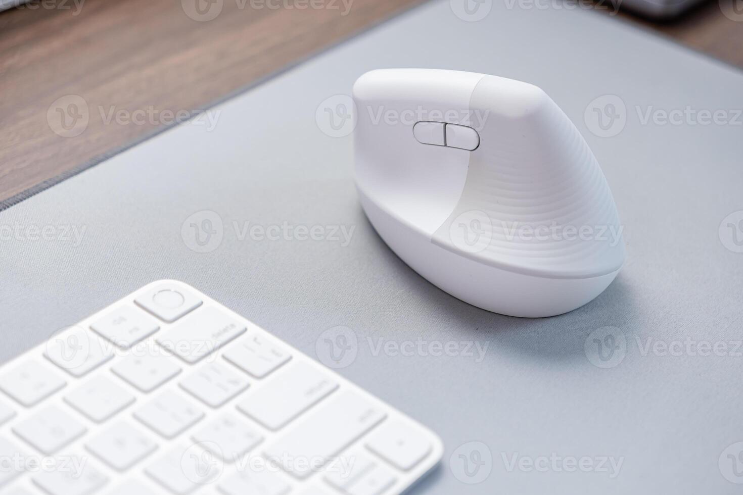 Modern white ergonomic vertical computer mouse sits on a grey desk mat next to a keyboard, emphasizing comfortable workspace setup and digital user health photo