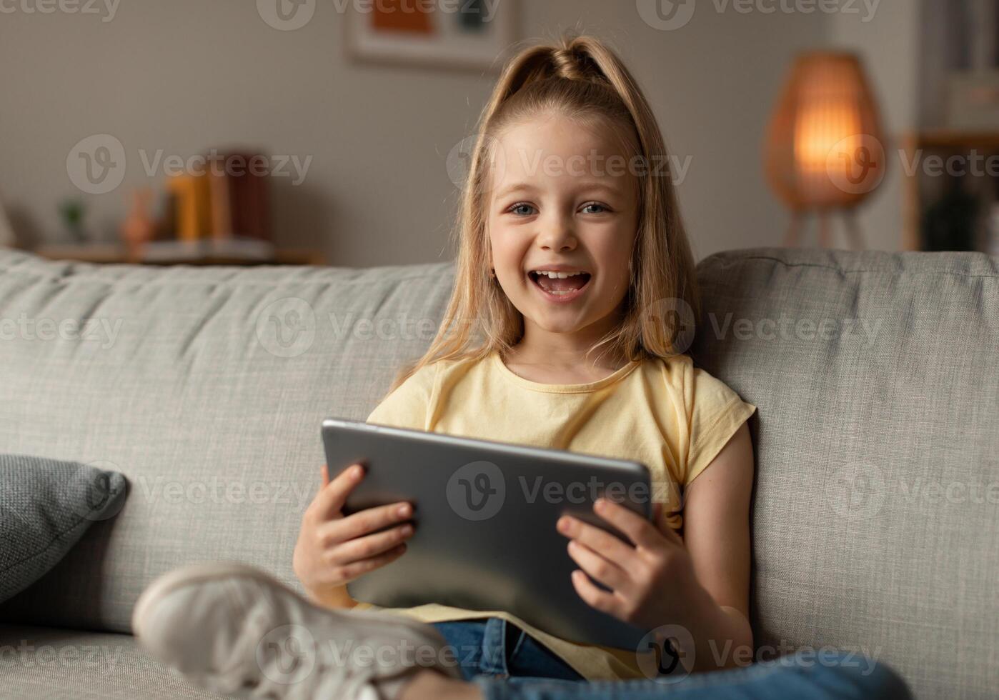Joyful Little Girl Laughing Using Digital Tablet Smiling To Camera Sitting On Sofa At Home. Kid Having Fun Browsing Internet And Playing Game Online On Computer. Children And Gadgets photo