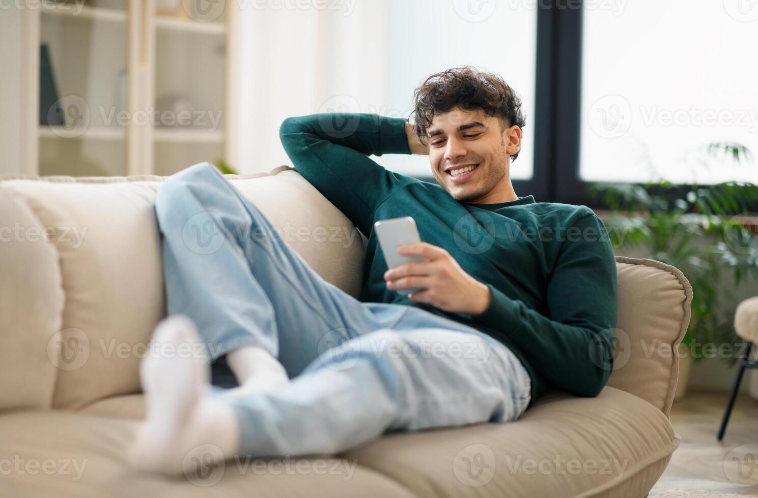 Mobile Offer. Smiling Middle Eastern Guy Browsing Internet On Smartphone, Communicating And Having Fun Online Lying On Couch At Home. Young Man Relaxing Using Gadget On Weekend. Selective Focus photo