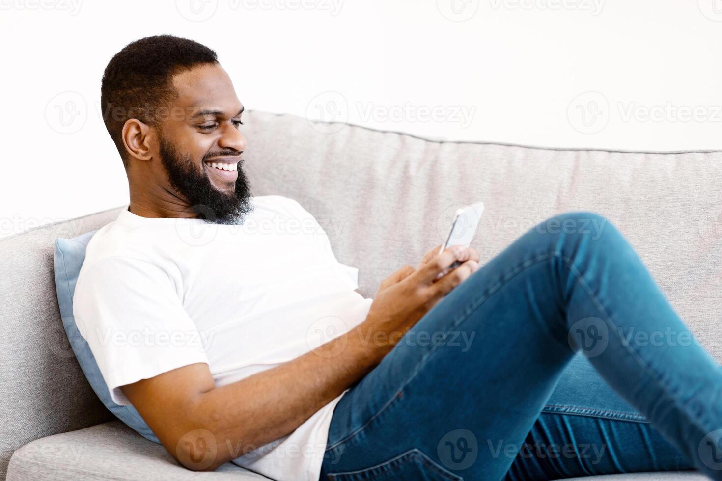 Mobile Communication. Black Guy Using Smartphone Browsing Internet And Texting Lying On Couch At Home, Side View. Mobile Phone User Networking Using Social Media Application. photo