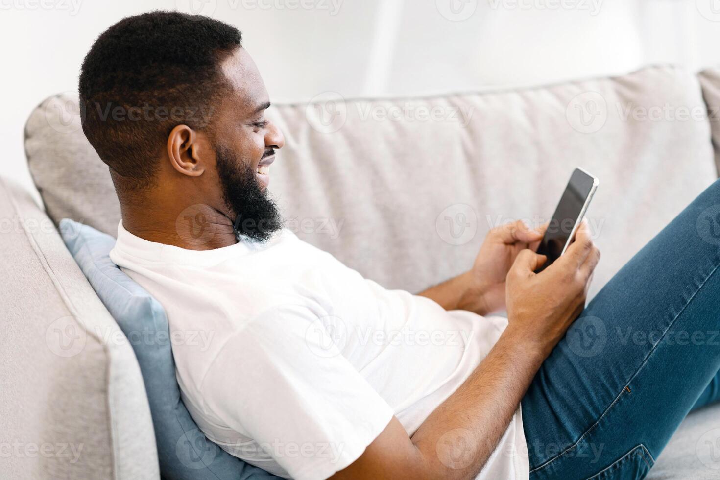 African American Man Using Smartphone Lying On Sofa At Home, Side View. Happy Mobile Phone User Browsing Internet And Texting, Networking In Social Media Indoors. Guy Testing New Application photo