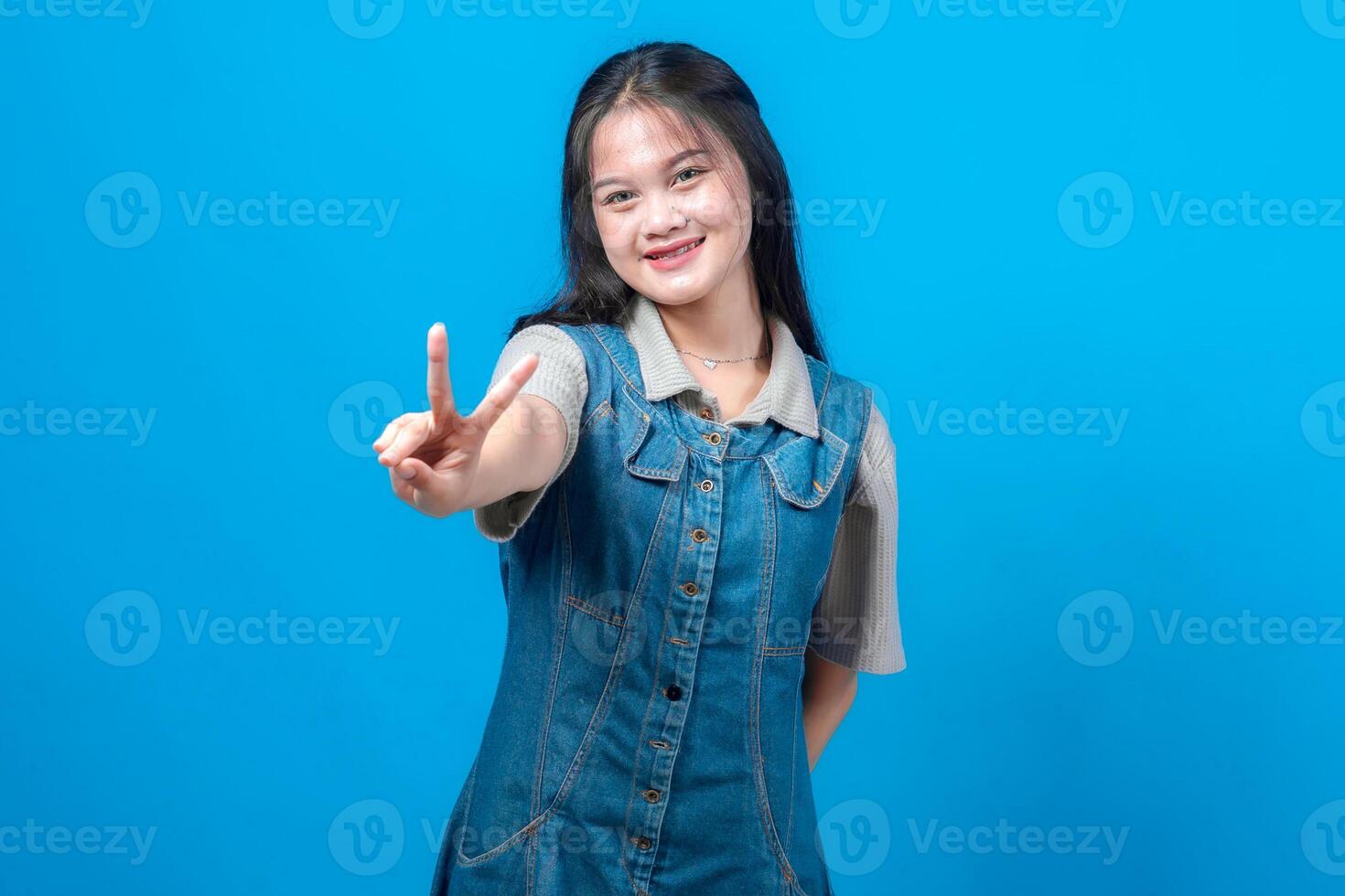joven asiático mujer sonriente y haciendo un paz firmar con su dedos extendido hacia el cámara. amigable, confidente expresión en casual mezclilla atuendo terminado azul fondo, estudio retrato. foto
