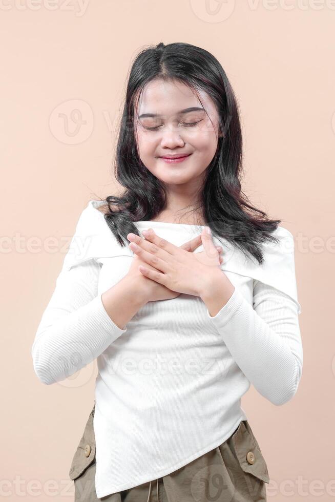 un joven asiático mujer cierra su ojos con un calma sonrisa y manos en su pecho, simbolizando gratitud, consciencia, y positivo emocional balance. foto