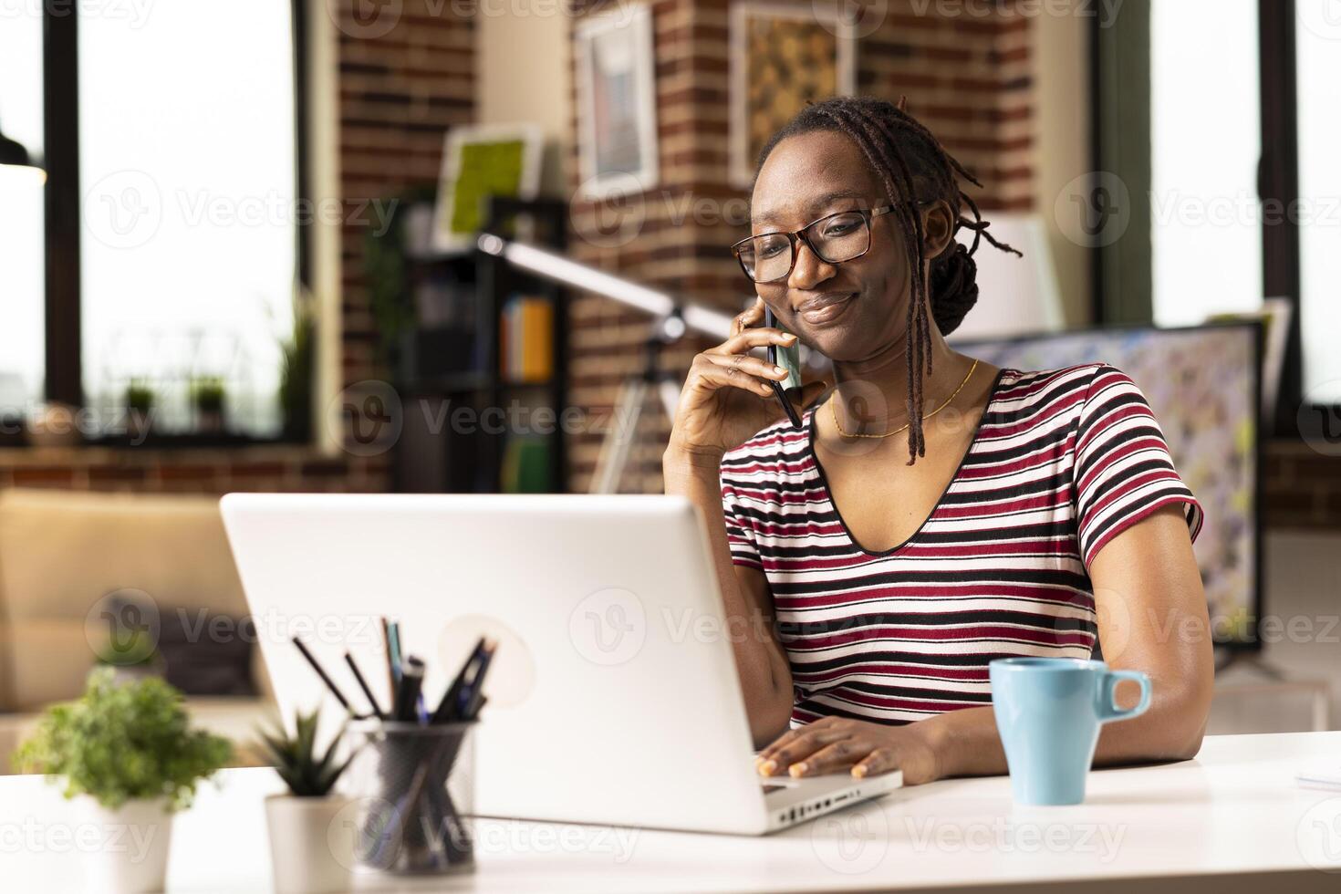 Self employed woman sits at desk, multitasking, speaks with clients on phone and updates data on laptop. Smiling female freelancer uses mobile device while working from home on personal computer. photo