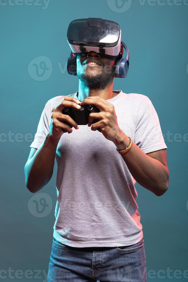 Young man playing a game with VR headset and controller, fully immersed in futuristic simulation. Male model with wireless headphones, holding a gamepad and enjoying a virtual reality interface. photo