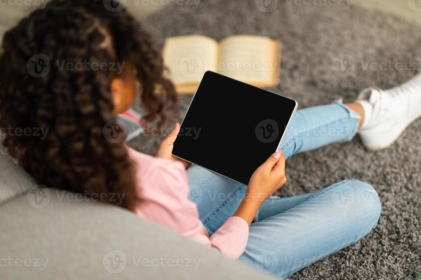 E-learning concept. African american schoolgirl using digital tablet with empty screen, surfing useful website or watching online lesson while sitting on floor with books. Mockup for your design photo