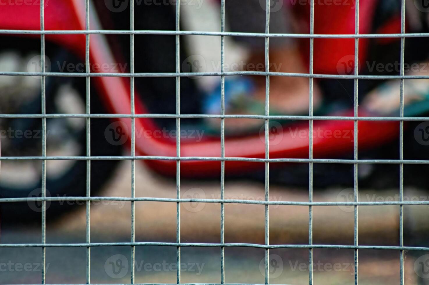 Close up of a metal wire fence with a blurred red vehicle and a wheel in the background, creating a sense of confinement or security, suitable for urban or industrial themes. photo
