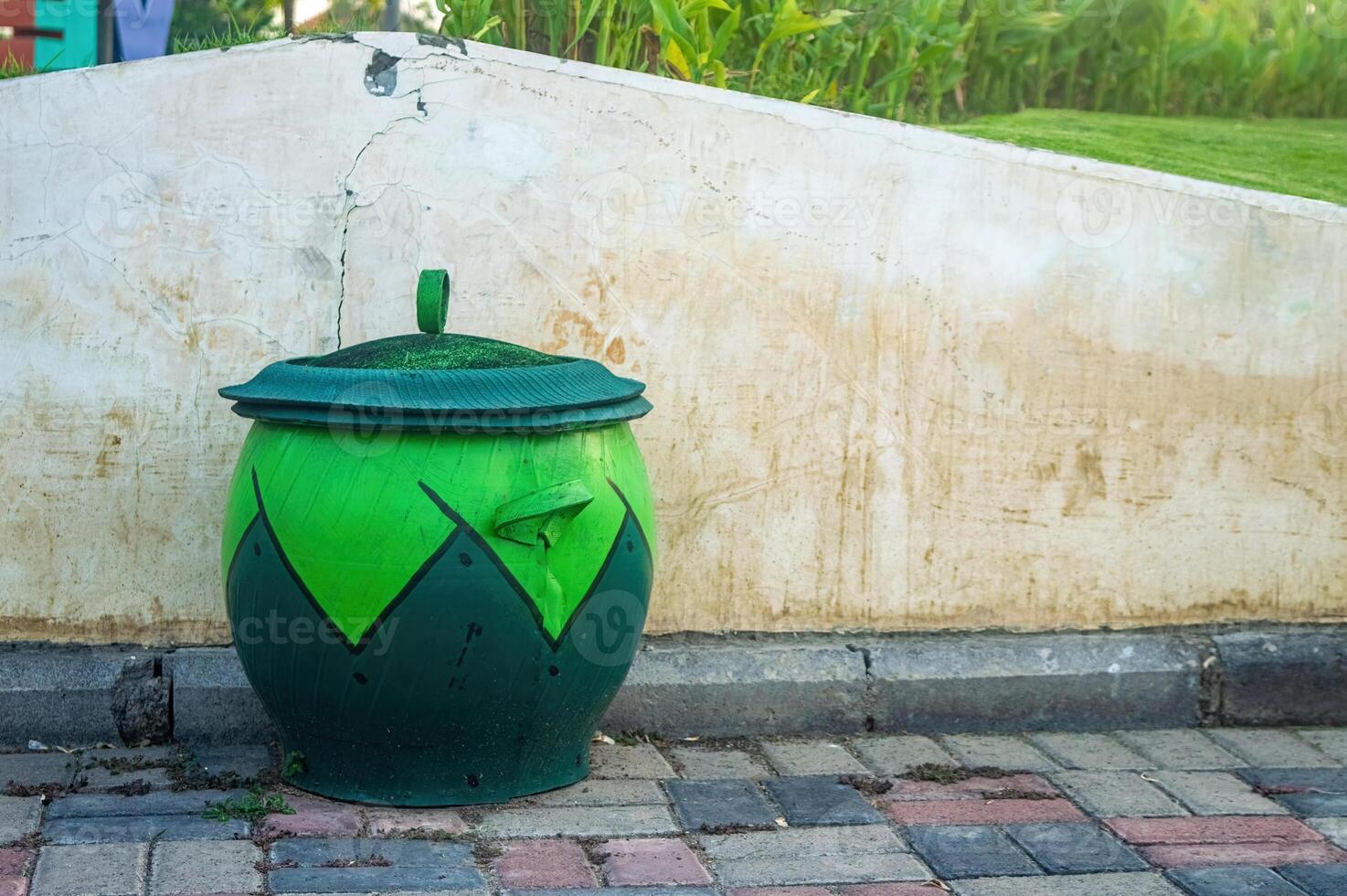 A green trash can with a lid sits on a brick sidewalk against a weathered wall. The trash can features a dark green geometric design, adding a touch of color to the urban setting. photo