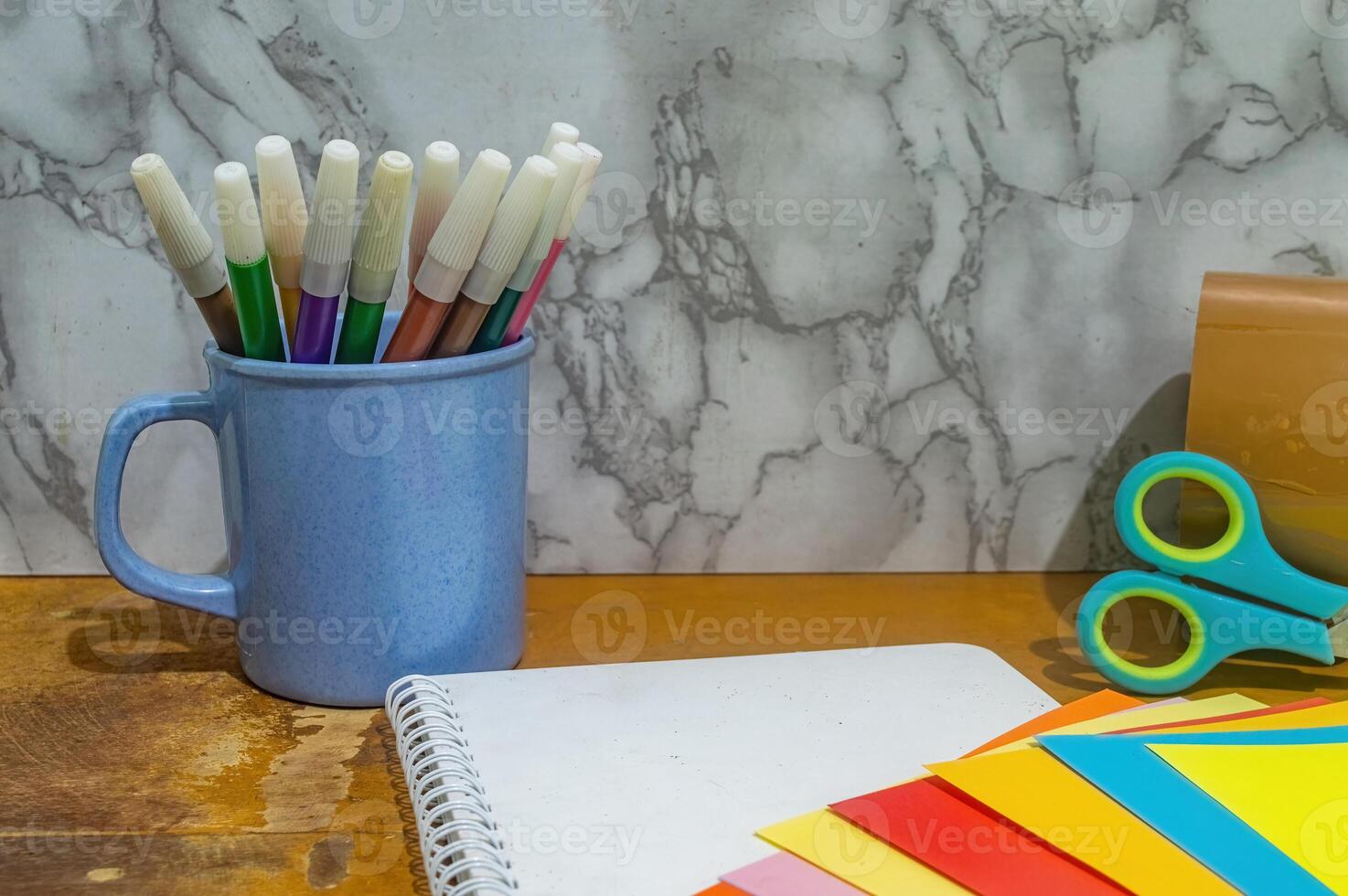 Close up shot displays a blue mug filled with colorful markers, a blank notebook, scissors, tape, and assorted color papers on a wooden table against a marble backdrop. Diy concept photo