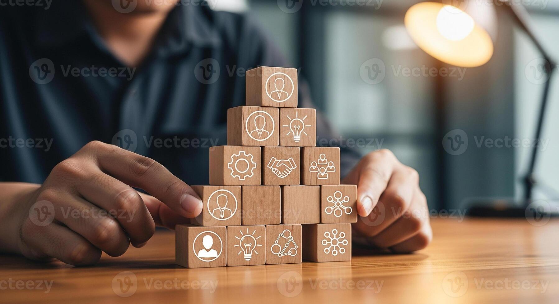 Person building a pyramid of wooden blocks with icons representing teamwork, strategy, and ideas in a modern office setting, symbolizing business growth and collaboration photo