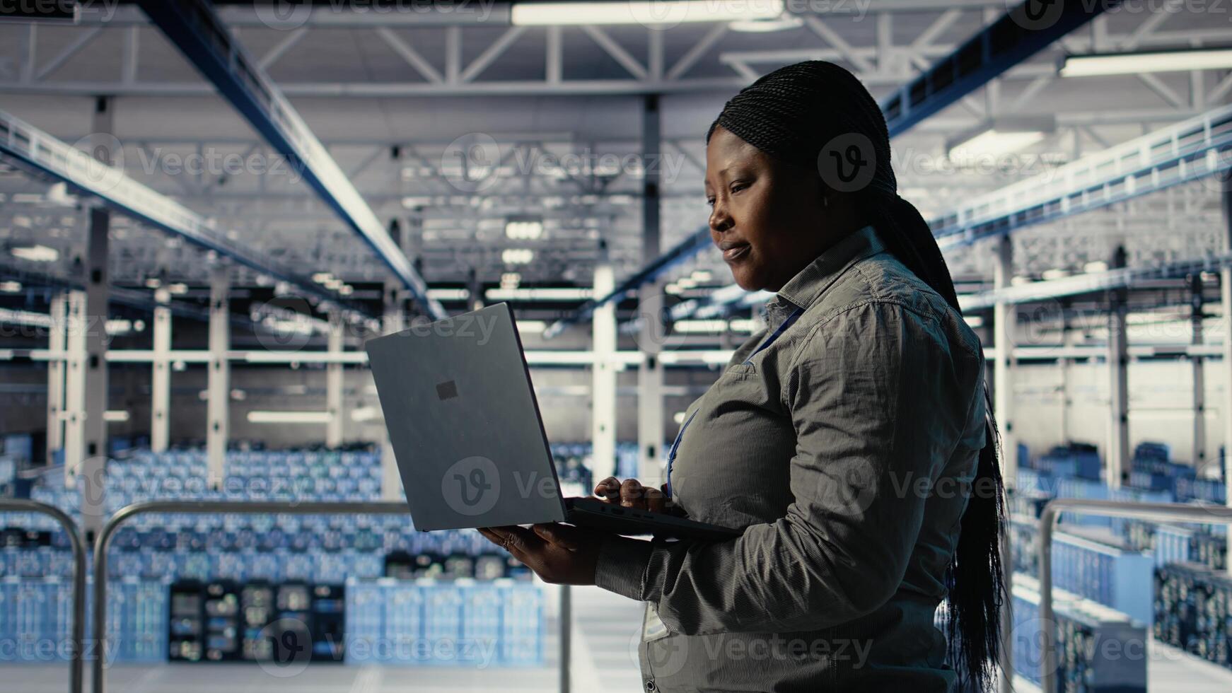 IT expert monitors server farm activity using laptop while walking in facility. African american woman in data center reviews rigs tests results to ensure systems are working correctly, camera A photo