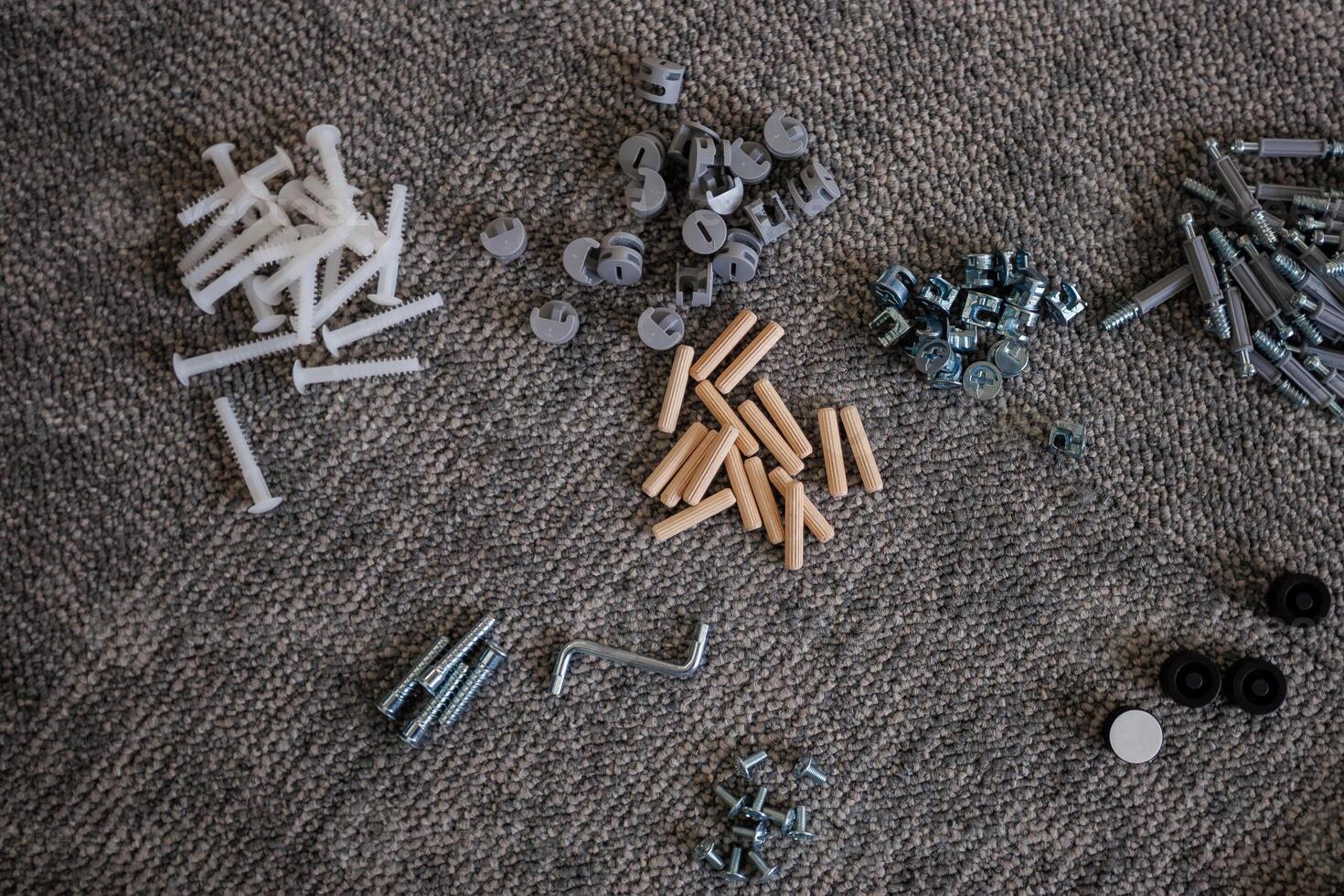 An assortment of hardware components, including screws, dowels, hex nuts, and an Allen wrench, neatly arranged on a textured carpet surface. photo
