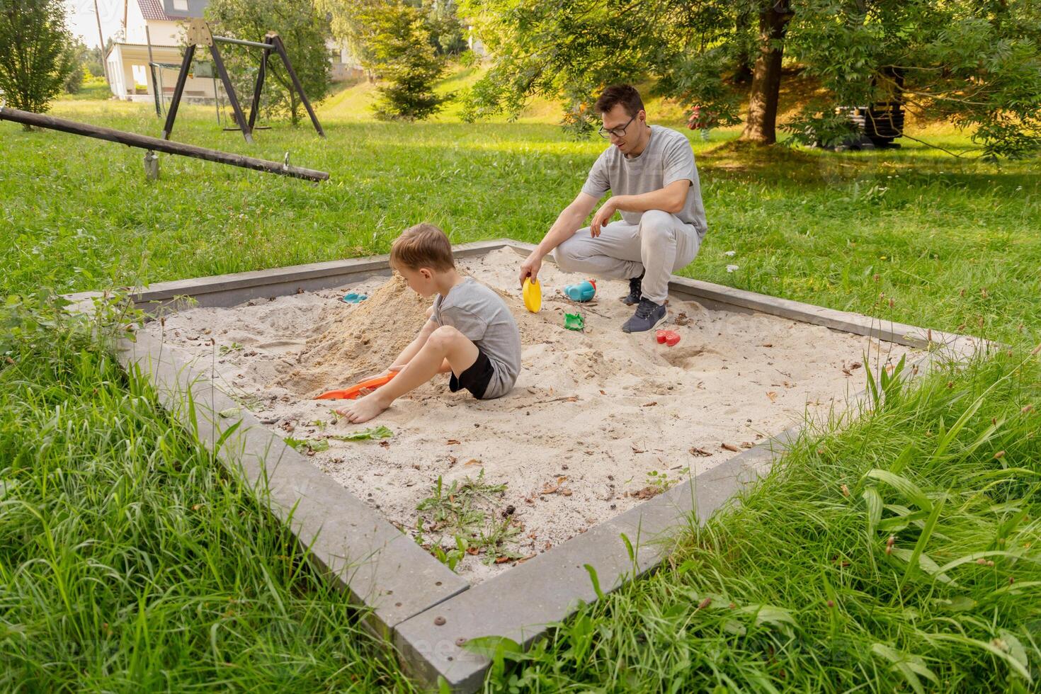 A father and his young child enjoy quality time together at a sandbox in a serene playground setting. Lush greenery surrounds them as they play and create memories photo