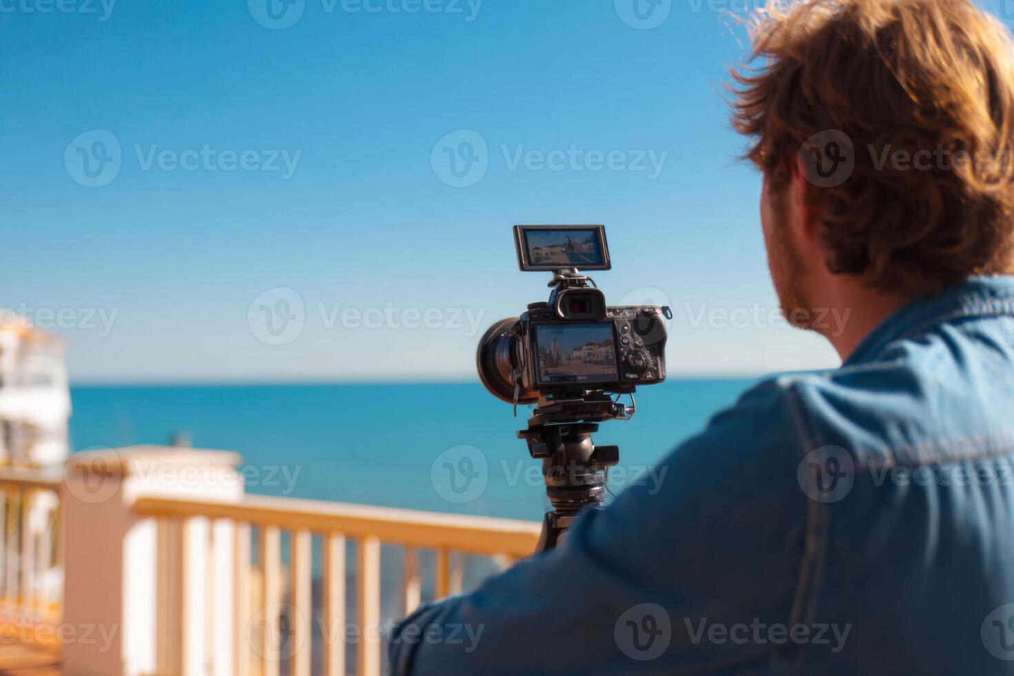 A person is using a camera on a tripod to take pictures of the ocean from a balcony. The clear blue sky and calm water enhance the serene atmosphere of the location during sunset photo