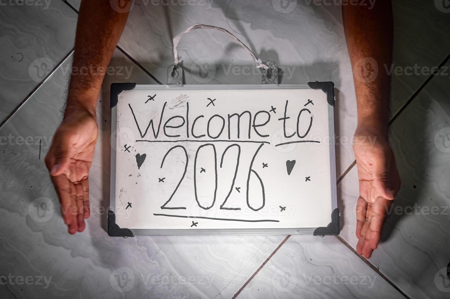 Whiteboard with Welcome to 2026 handwritten in playful style, framed by two open hands on tiled floor with heart and X doodles symbolizing new year warmth and fresh beginnings. photo