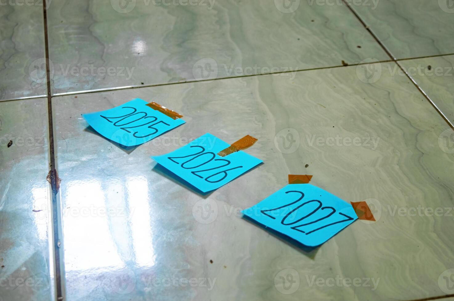 Three blue sticky notes labeled 2025, 2026, and 2027 taped to tiled floor symbolizing yearly progress, future planning, and timeline goals in a minimalist setup. photo
