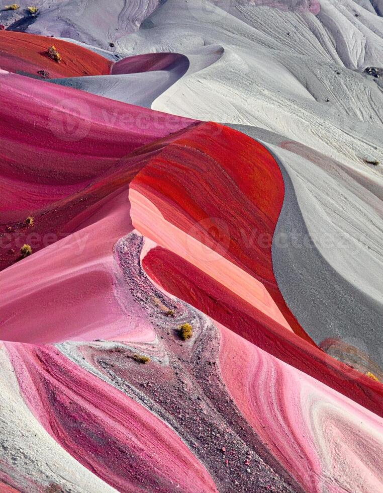 Abstract landscape of vibrant, swirling hills. Close-up view of textured geological formations in various shades of pink, red, gray, and white. Intricate patterns and curves dominate the image, with small vegetation sprinkled across the landscape photo