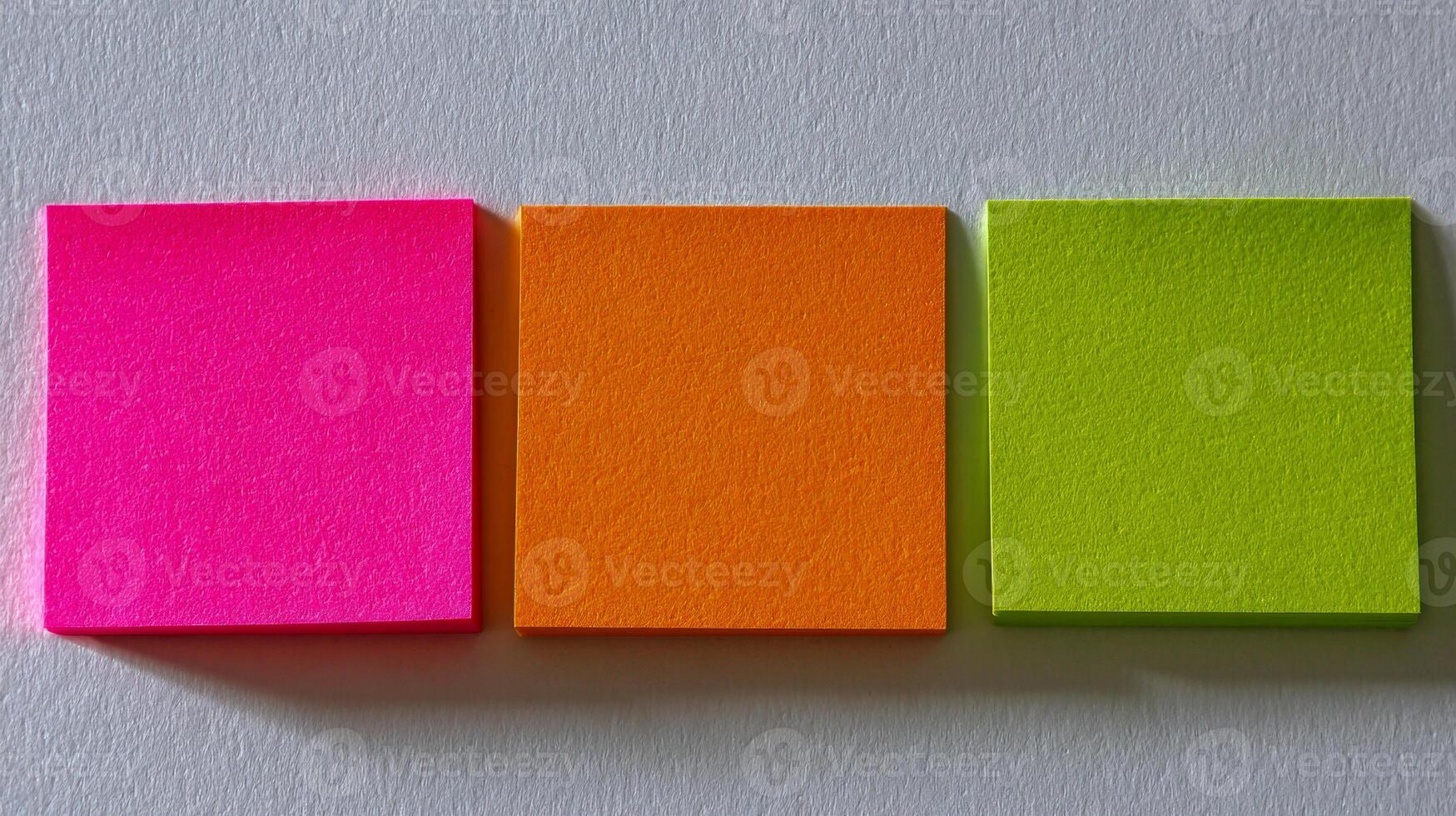 Bright neon pink, orange, and green sticky notes aligned horizontally on a plain light background with soft shadows adding depth and contrast to the composition photo