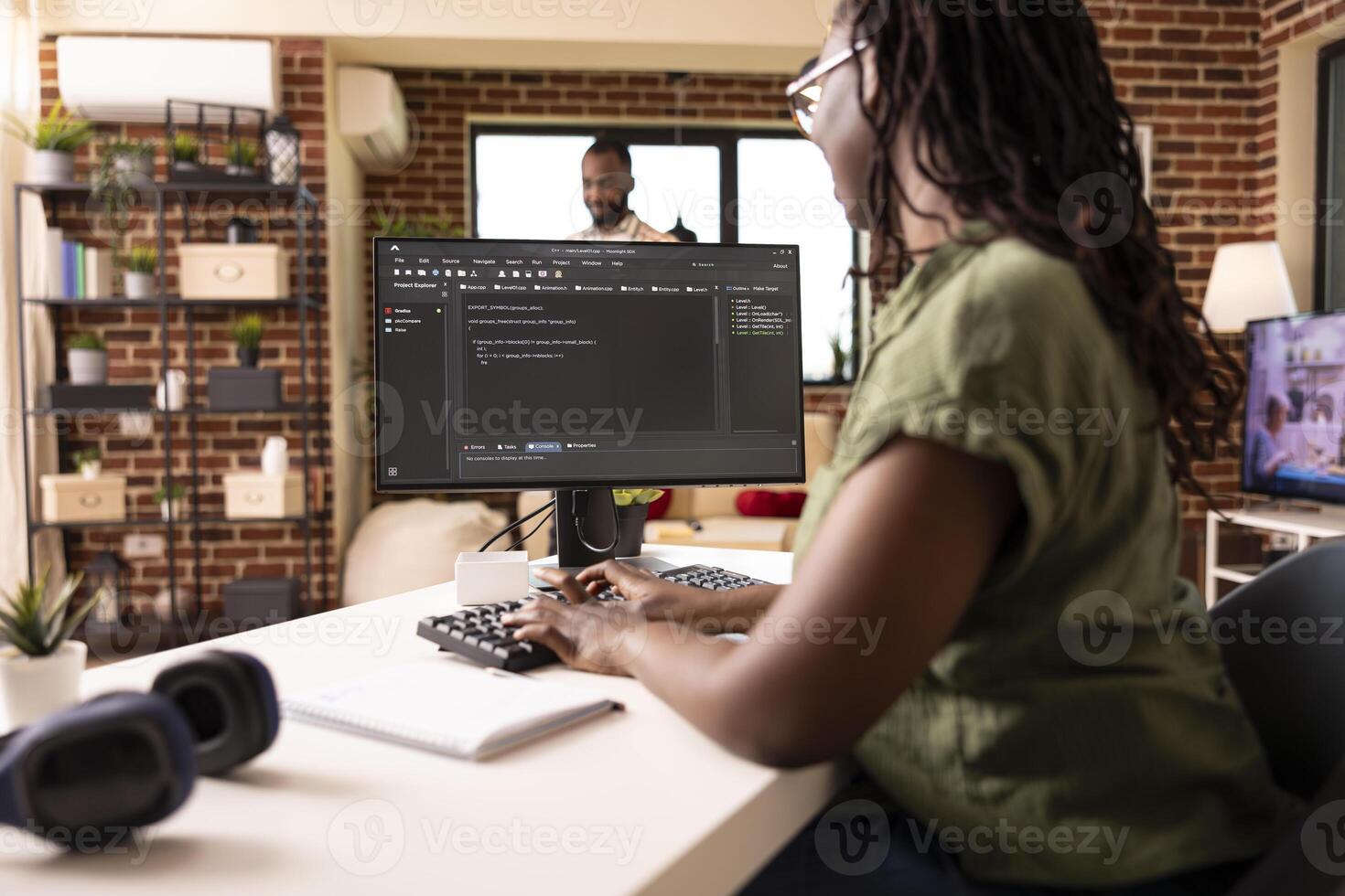 Remote female programmer works from cozy home desk, coding for startup project on desktop pc. Tech savvy black woman managing web development tasks on computer in brick wall apartment. photo