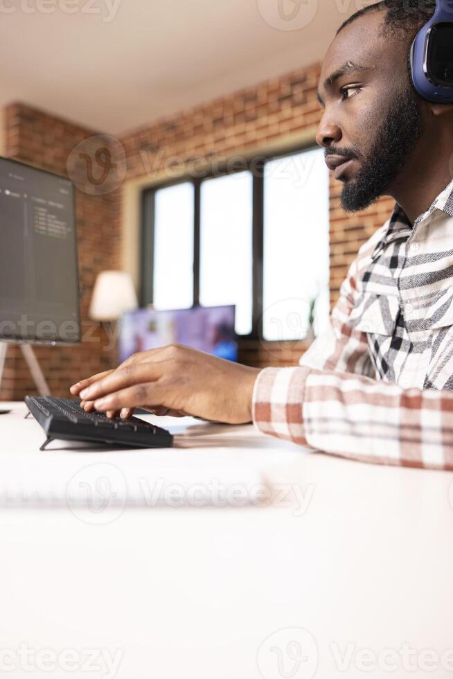 Black male developer wears headphones while coding from home, reviewing programming scripts on computer. Remote IT freelancer working on software solutions in modern apartment. photo