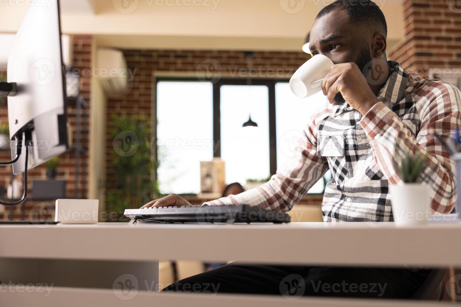 African american man entrepreneur sips tea while analyzing business data and reading digital content on desktop pc at home. Male copywriter drinking coffee and reviewing web articles on computer. photo