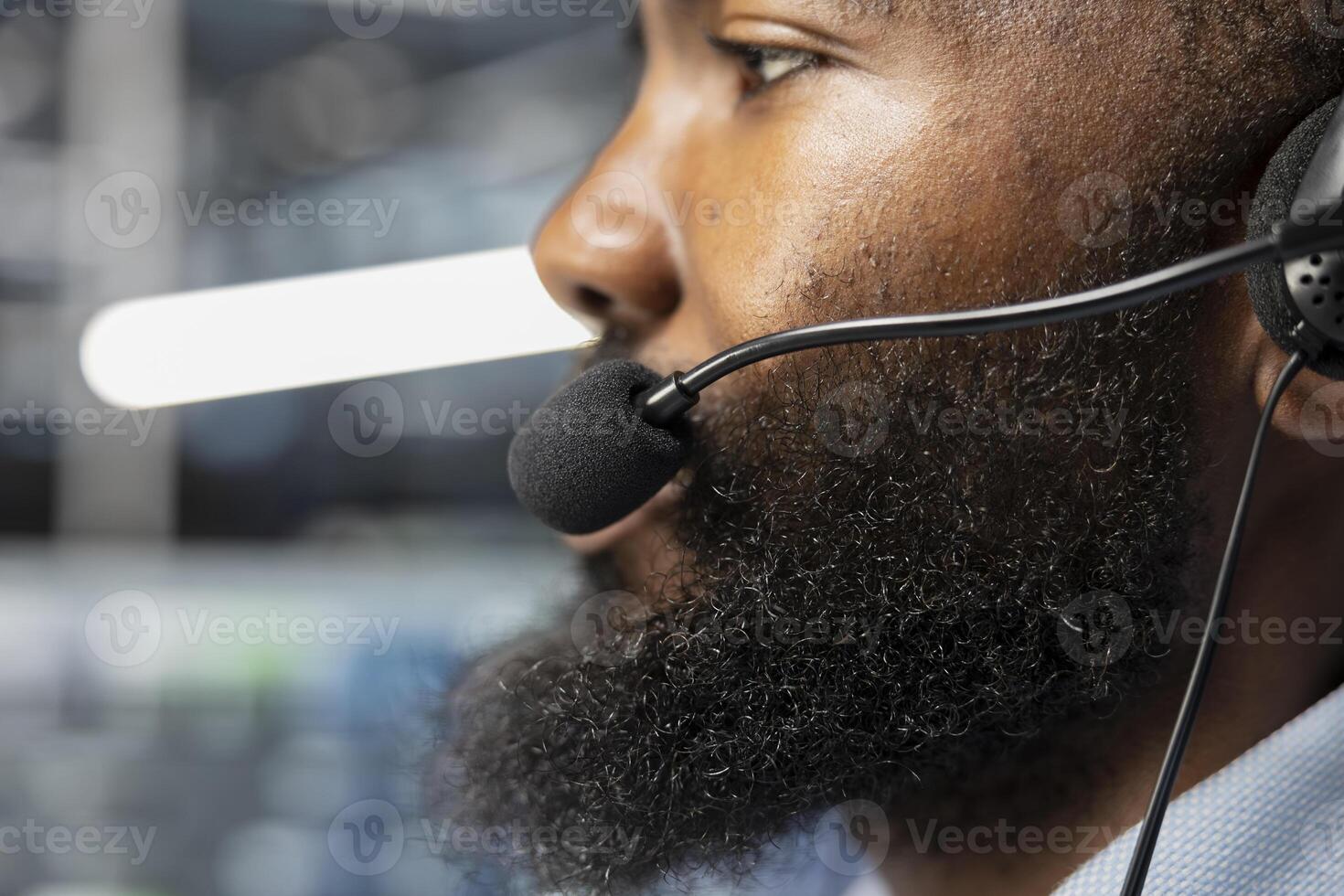 Close up of data center worker in remote call providing customer support, troubleshooting damages. African american man in server hub talking with clients, using microphone photo