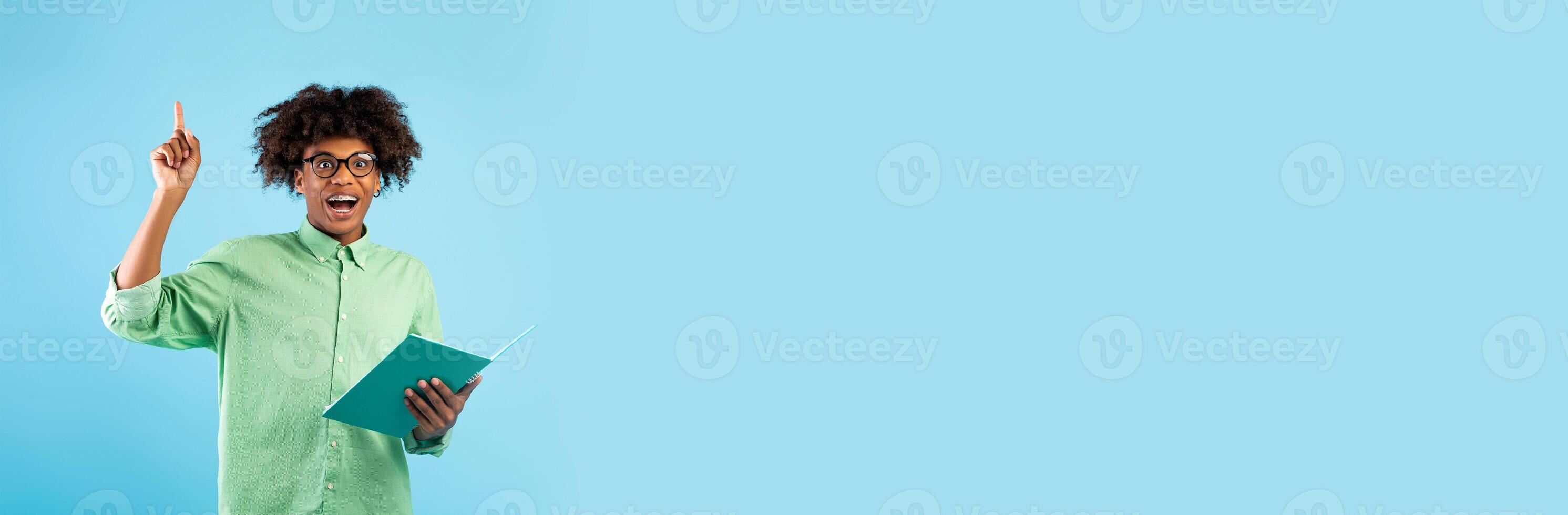 A student holds a book while raising a finger in a thoughtful gesture, showcasing an expression of inspiration. The bright background provides ample space for text or messaging. photo