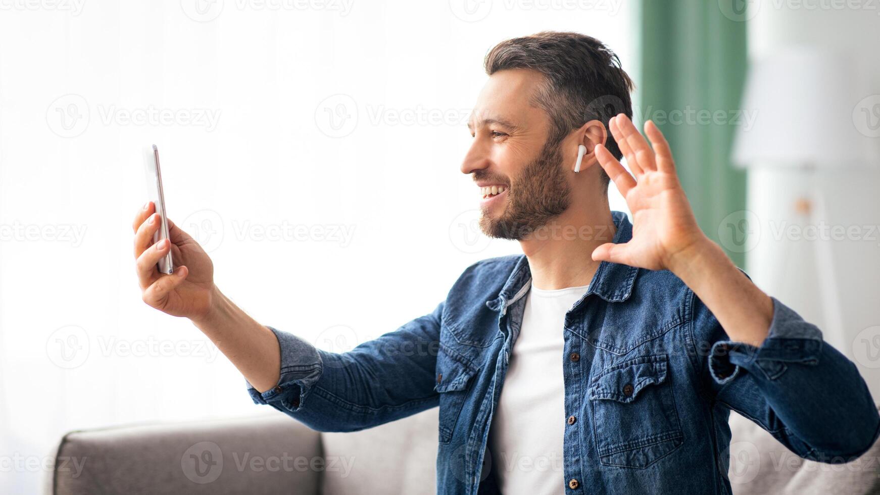 Cheerful bearded man waving at mobile phone while resting on couch at home, side view, copy space. Happy middle-aged man having conference with friends or relatives, using smartphone photo
