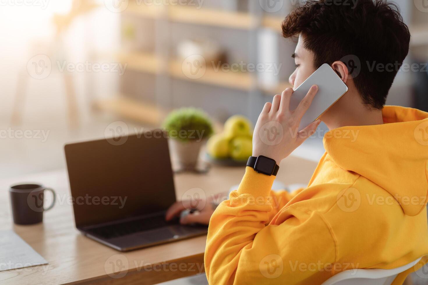 Over the shoulder view of asian man having conversation on mobile phone, using laptop with blank screen, copy space. Male freelancer talking with clients on smartphone while using pc at desk photo