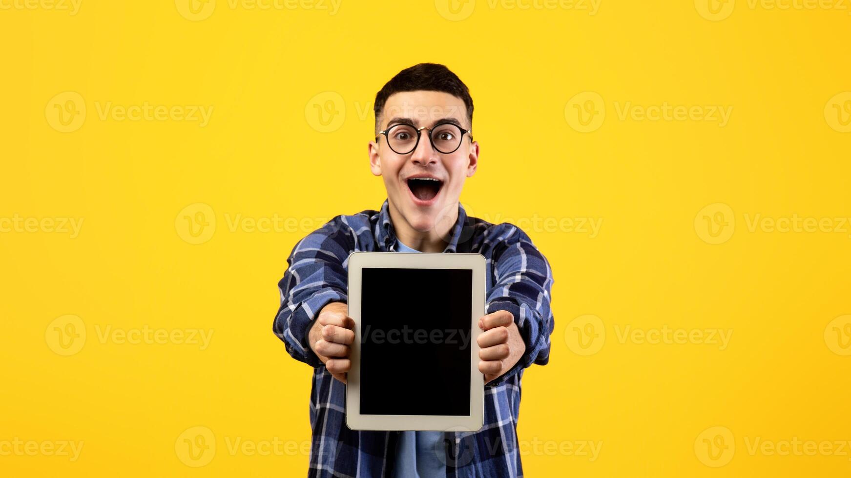 Overjoyed millennial man showing tablet computer with mockup for website or app design on orange studio background. Excited young guy with touch pad, space for your ad photo