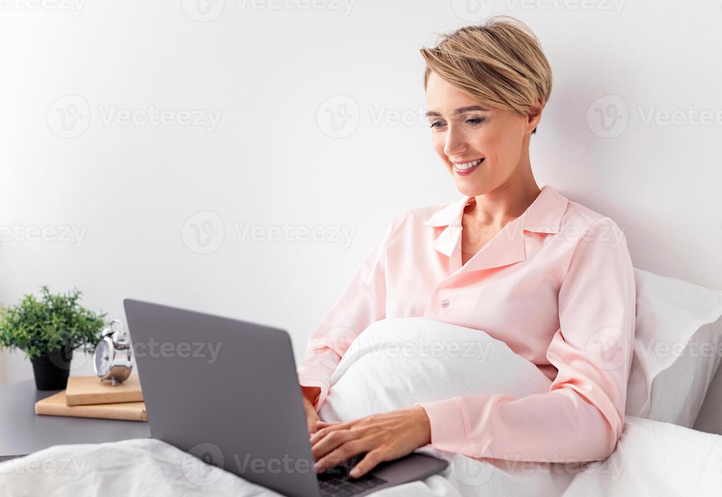 Portrait Of Smiling Beautiful Middle Aged Woman In Pink Sleepwear Sitting On Bed Under Blanket At Home, Using Computer Typing On Laptop Keyboard, Browsing Internet, Working Or Resting photo